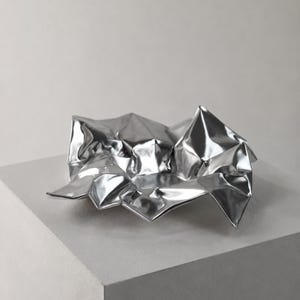 May include: A silver-colored bowl with a highly reflective, metallic surface. The bowl features an irregular, crumpled, and faceted shape with sharp, angular folds and deep creases that create a complex pattern of light and dark shadows.