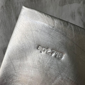May include: A rectangular silver-toned metal object with a brushed finish. The surface features the stamped text "apt.716" in a lowercase sans-serif font. The metal shows visible diagonal scratch marks across its entire surface, creating a textured appearance.