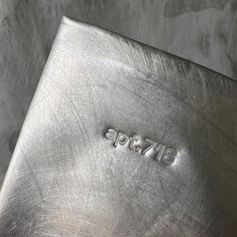 May include: A rectangular silver-toned metal object with a brushed finish. The surface features the stamped text "apt.716" in a lowercase sans-serif font. The metal shows visible diagonal scratch marks across its entire surface, creating a textured appearance.