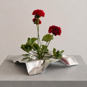 May include: A vibrant red geranium plant with green leaves is arranged in a silver-colored, metallic, crinkled, star-shaped vessel. The plant features multiple stems with clusters of deep red flowers and broad, textured green foliage.