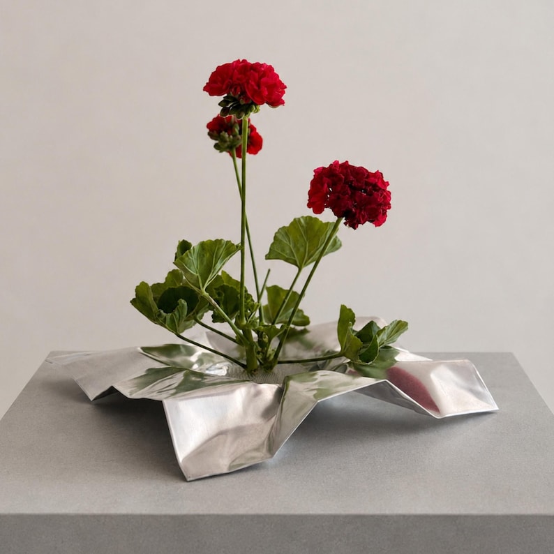 May include: A vibrant red geranium plant with green leaves is arranged in a silver-colored, metallic, crinkled, star-shaped vessel. The plant features multiple stems with clusters of deep red flowers and broad, textured green foliage.