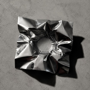 May include: A square, silver-colored metallic bowl with a highly reflective, crinkled surface that mimics the appearance of folded foil. The center of the bowl features a circular, textured base with a dense pattern of small, raised dots.