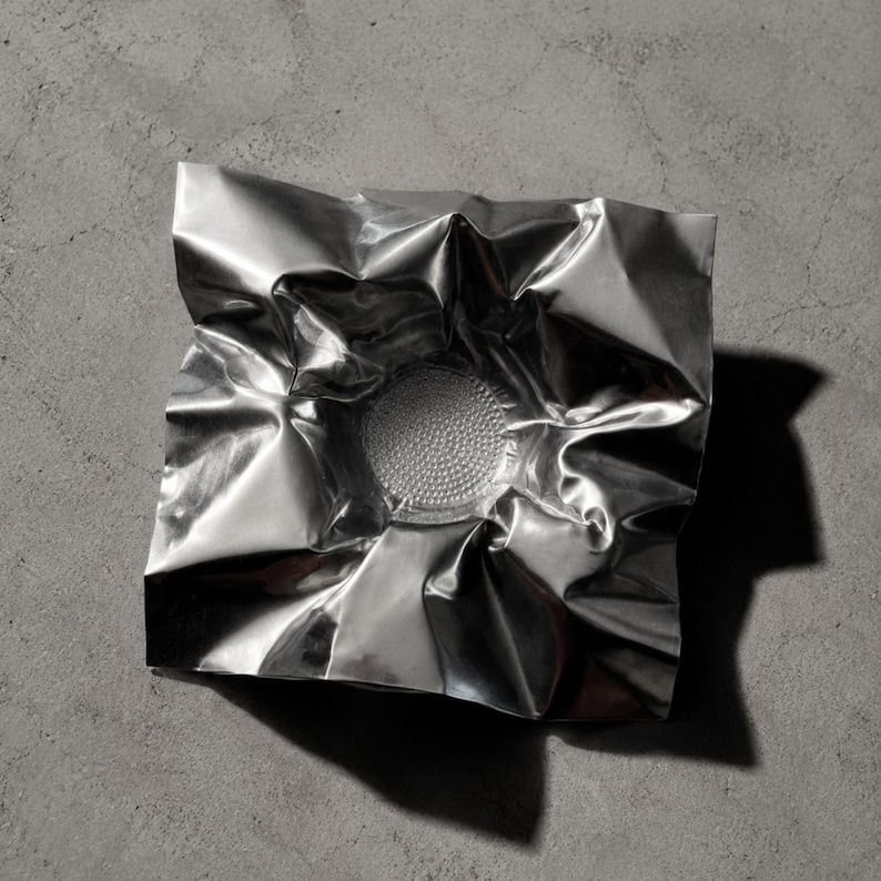 May include: A square, silver-colored metallic bowl with a highly reflective, crinkled surface that mimics the appearance of folded foil. The center of the bowl features a circular, textured base with a dense pattern of small, raised dots.