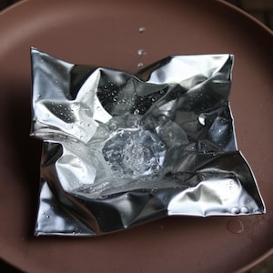 May include: A square sheet of crinkled, reflective silver foil shaped into a shallow basin. The center of the foil holds a small pool of clear water, with several distinct water droplets scattered across the metallic, uneven surface.