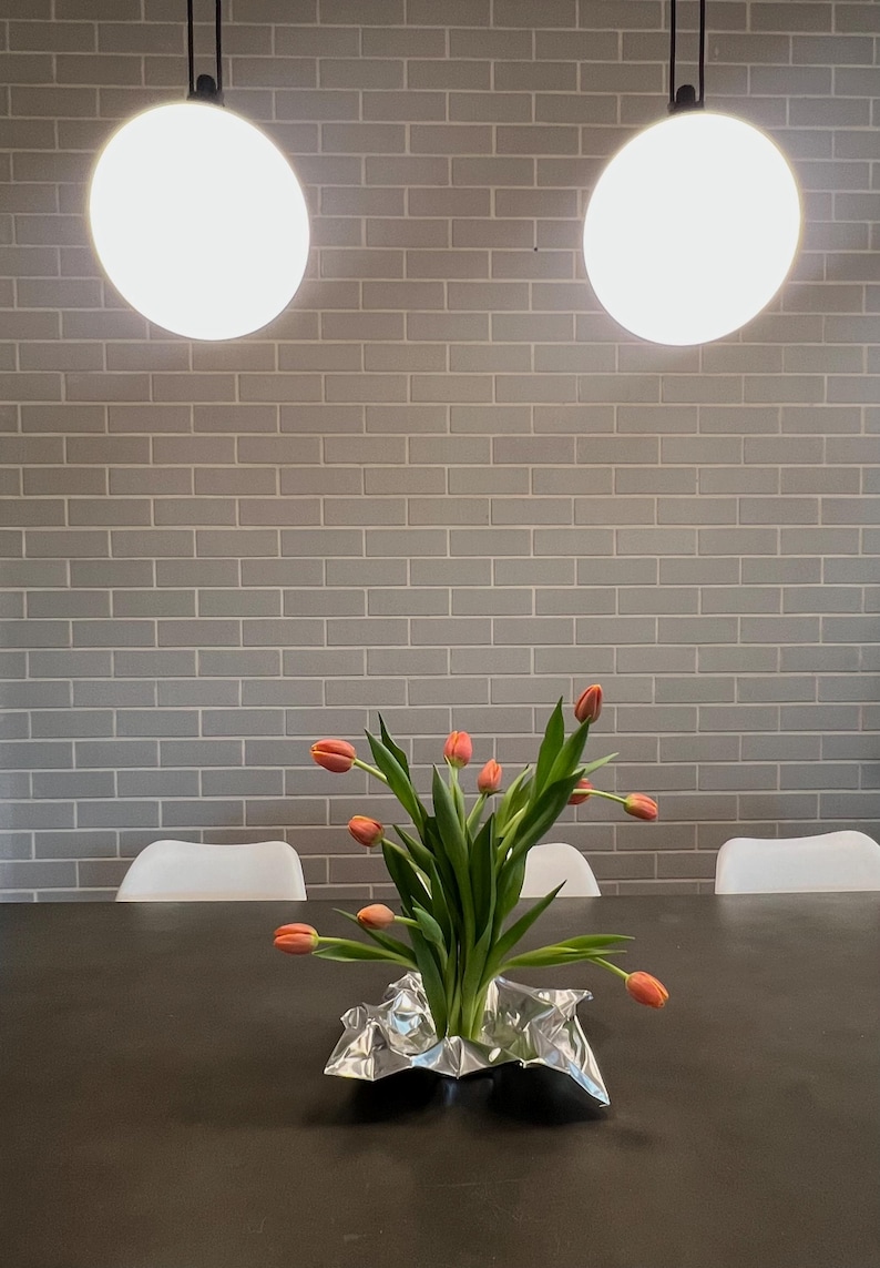 May include: A bouquet of orange tulips with green stems and leaves arranged in a crinkled silver metallic foil base. The flowers are positioned on a dark surface, with the stems gathered together at the center of the reflective silver material.