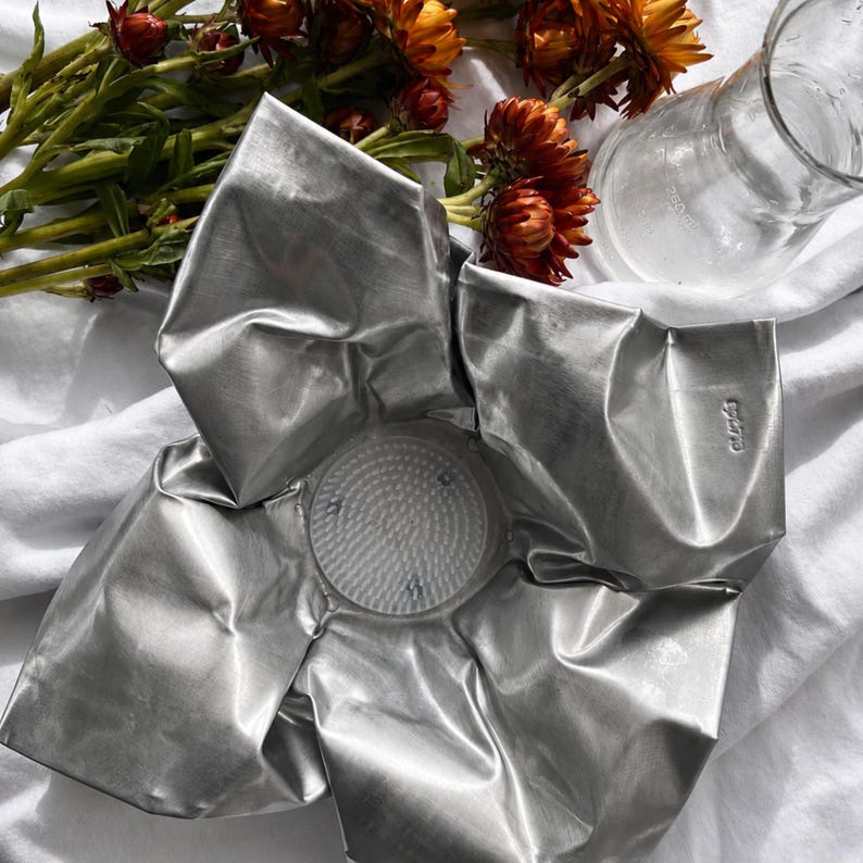 May include: A silver-colored, metallic flower-shaped bowl with five large, folded petals. The center features a circular, textured base with a grid pattern. The surface of the metal has a crinkled, reflective finish with visible folds and creases.
