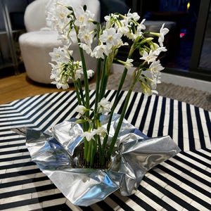 May include: A cluster of white narcissus flowers with green stems arranged in a circular metal floral frog. The base of the arrangement is wrapped in a reflective, crinkled silver metallic sheet that flares outward to create a sculptural display.