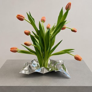May include: A bouquet of orange tulips with green stems and leaves arranged in a reflective, silver-colored metallic vase that features a crinkled, crumpled texture resembling foil, creating a modern and industrial aesthetic for floral displays.