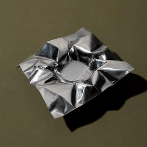 May include: A square, silver-colored metallic sheet with a crinkled, reflective surface. The center of the sheet is depressed into a bowl-like shape, featuring a circular, textured base with numerous small, upright, needle-like protrusions.