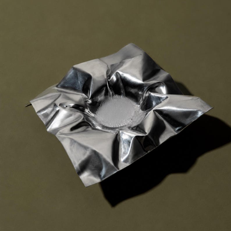 May include: A square, silver-colored metallic sheet with a crinkled, reflective surface. The center of the sheet is depressed into a bowl-like shape, featuring a circular, textured base with numerous small, upright, needle-like protrusions.
