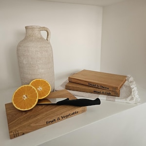 May include: Wooden cutting boards with engraved labels, including "Fruit & Vegetable", "Bread & Cheese", and "Meat & Fish". A black-handled knife and sliced oranges are also present. A beige jug and a white cloth complete the kitchen scene.