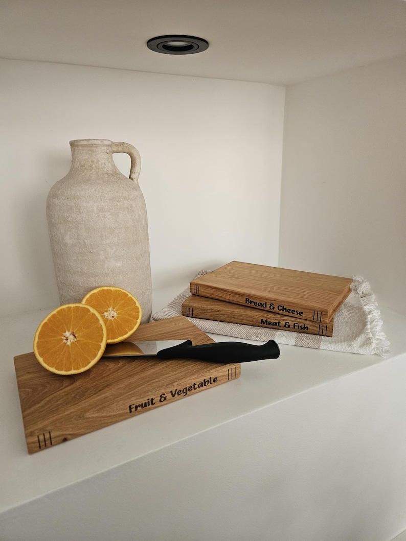 May include: Wooden cutting boards with engraved labels, including "Fruit & Vegetable", "Bread & Cheese", and "Meat & Fish". A black-handled knife and sliced oranges are also present. A beige jug and a white cloth complete the kitchen scene.