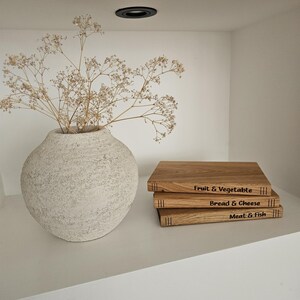 May include: A textured, off-white vase holds dried flowers. Stacked wooden cutting boards with engraved labels: "Fruit & Vegetable," "Bread & Cheese," and "Meat & Fish" sit on a white shelf. The scene is illuminated by a recessed ceiling light.
