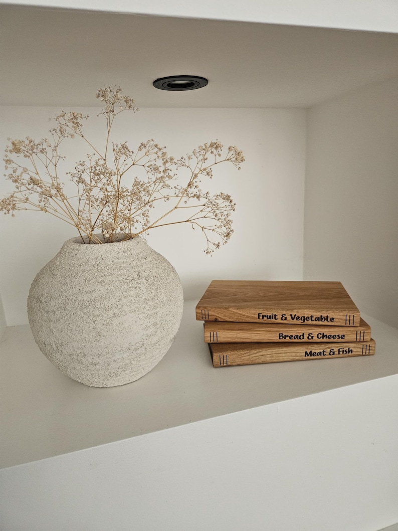May include: A textured, off-white vase holds dried flowers. Stacked wooden cutting boards with engraved labels: "Fruit & Vegetable," "Bread & Cheese," and "Meat & Fish" sit on a white shelf. The scene is illuminated by a recessed ceiling light.