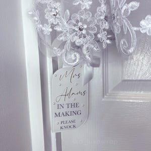 May include: A white door hanger with the text "Mrs Adams IN THE MAKING PLEASE KNOCK" in black script. The hanger is decorated with small, silver-coloured embellishments. White floral lace fabric is visible in the background.