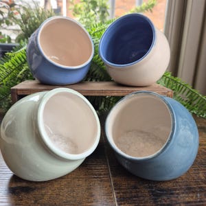 May include: Four ceramic salt cellars in shades of blue, green, and beige. Each has a rounded shape with a wide opening, and some are filled with salt. The cellars are arranged on a wooden surface.