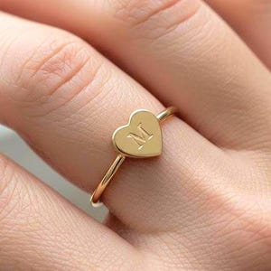 May include: A gold-colored ring featuring a heart-shaped design with the letter "M" engraved on the heart. The ring is simple and elegant, with a thin band. The ring is worn on a finger.