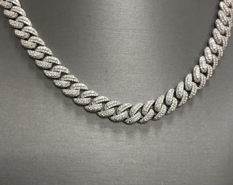Op de afbeelding: Een zilverkleurige, met diamanten bezette Cuban link ketting. De ketting wordt getoond op een donkergrijze mannequin. De schakels van de ketting zijn nauw met elkaar verbonden, wat een dikke, luxueuze uitstraling creëert. De diamanten schitteren en reflecteren licht.