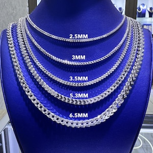 May include: A display of silver chain necklaces in varying thicknesses, labeled 2.5MM, 3MM, 3.5MM, 5.3MM, and 6.5MM. The chains are arranged on a blue surface, showcasing their different designs and sizes.