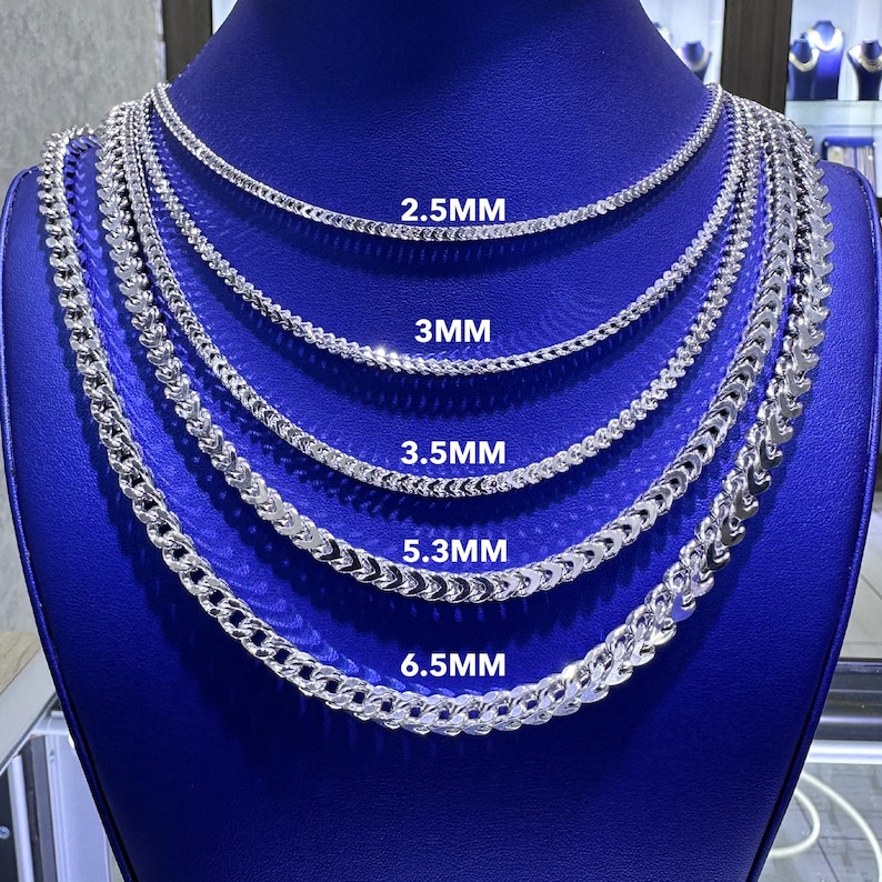 May include: A display of silver chain necklaces in varying thicknesses, labeled 2.5MM, 3MM, 3.5MM, 5.3MM, and 6.5MM. The chains are arranged on a blue surface, showcasing their different designs and sizes.