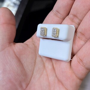 以下が含まれることがあります： 長方形のスタッドピアス。ゴールドカラーのフレームと小さなクリアストーンが特徴です。白いジュエリーカードに展示されており、人の手で持たれています。ジュエリーアイテムです。