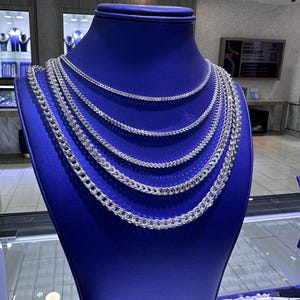 May include: A blue jewelry display bust showcases multiple silver chain necklaces. The necklaces vary in thickness and style, creating a layered effect. The display is in a jewelry store setting, with additional jewelry visible in the background.