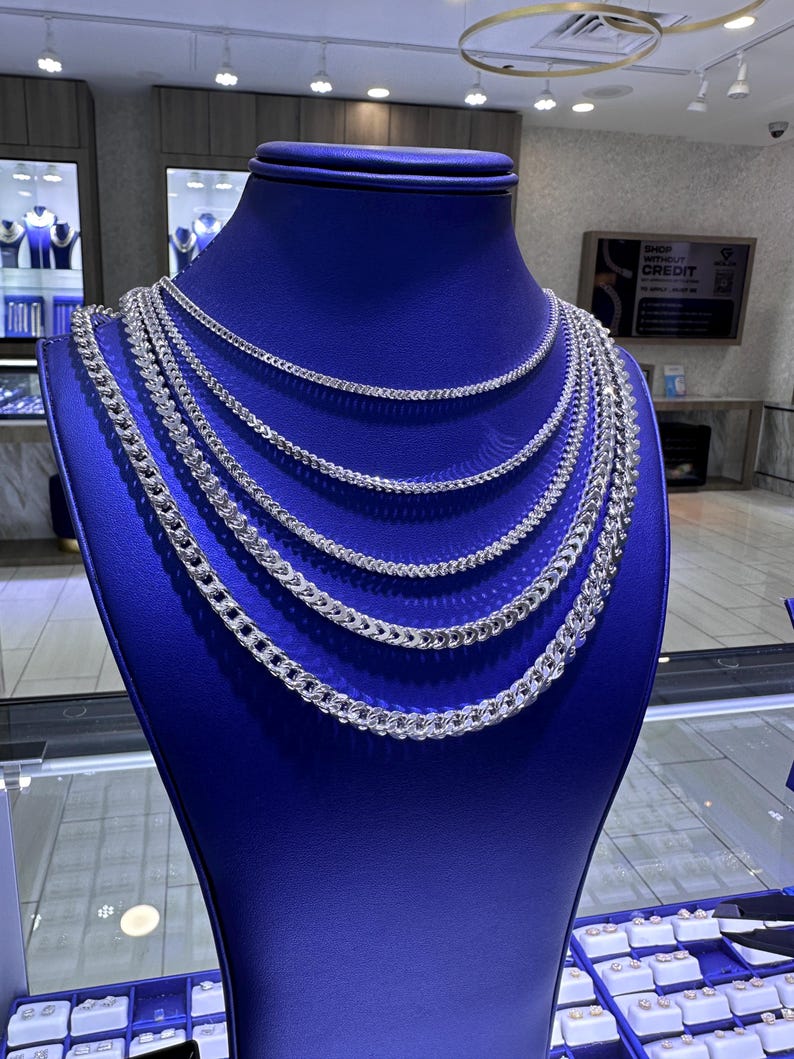 May include: A blue jewelry display bust showcases multiple silver chain necklaces. The necklaces vary in thickness and style, creating a layered effect. The display is in a jewelry store setting, with additional jewelry visible in the background.