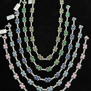 May include: A collection of silver necklaces with flower-shaped pendants in various colors, including pink, blue, and green. Each necklace is approximately 16 inches long, displayed against a black background.