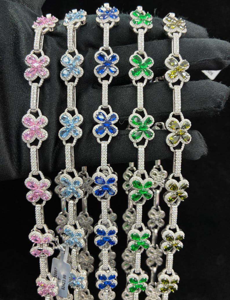 May include: A collection of silver bracelets, each featuring a floral design with various colored gemstones. The bracelets are adorned with pink, light blue, dark blue, green, and olive-green flower-shaped stones, and are connected by a line of small, clear stones.