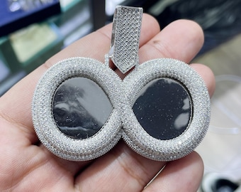 Puede incluir: Un colgante de plata en forma de infinito con dos ranuras redondas para fotos, cada una rodeada por un borde de pequeñas piedras de circonita cúbica transparentes.