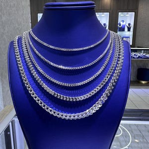 May include: A display of silver chain necklaces of varying thicknesses on a blue mannequin. The necklaces are arranged in layers, showcasing different styles of chain links. The background includes a jewelry store setting with display cases and lighting.