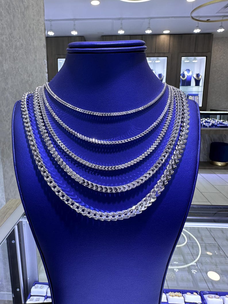 May include: A display of silver chain necklaces of varying thicknesses on a blue mannequin. The necklaces are arranged in layers, showcasing different styles of chain links. The background includes a jewelry store setting with display cases and lighting.