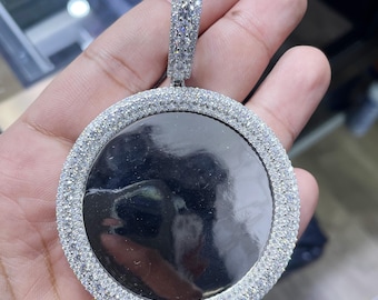 Puede incluir: Un colgante redondo de plata con un centro transparente y un borde de pequeñas piedras transparentes. El colgante está unido a una cadena de plata.