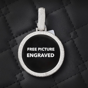 May include: A silver pendant with a circular, black centre. The pendant is edged with small, clear stones and has a bail also covered in stones. The text "FREE PICTURE ENGRAVED" is displayed in white on the black centre.