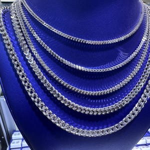 May include: A display of silver-colored chain necklaces of varying thicknesses. The chains are arranged on a blue display stand, showcasing different styles and sizes. The necklaces have a shiny, reflective surface.