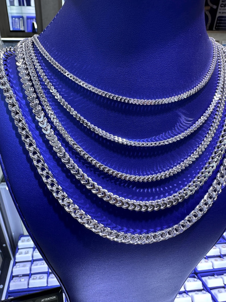 May include: A display of silver-colored chain necklaces of varying thicknesses. The chains are arranged on a blue display stand, showcasing different styles and sizes. The necklaces have a shiny, reflective surface.