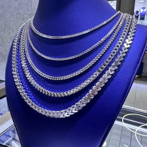 May include: A display of multiple silver chain necklaces on a blue mannequin. The necklaces vary in style and thickness, with a bright, reflective finish. The chains are arranged to showcase their different designs and lengths.