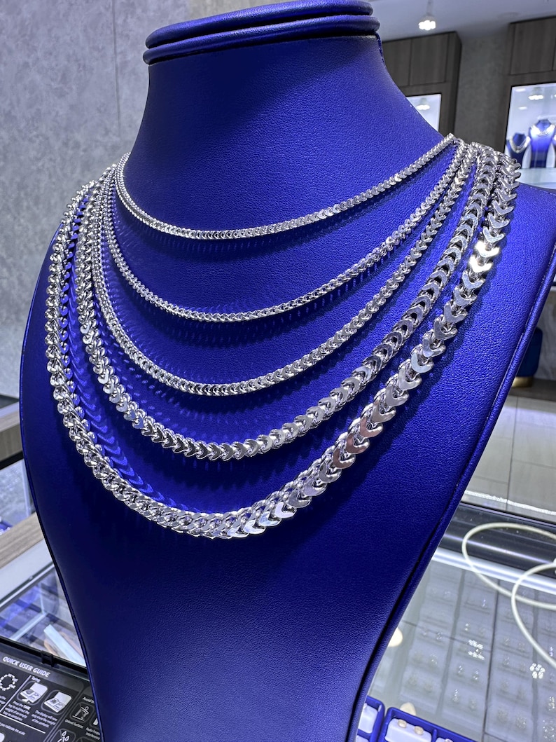 May include: A display of multiple silver chain necklaces on a blue mannequin. The necklaces vary in style and thickness, with a bright, reflective finish. The chains are arranged to showcase their different designs and lengths.