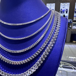 May include: A display of silver necklaces with various chain styles. The necklaces are arranged on a blue mannequin, showcasing different thicknesses and designs. The chains have a shiny, reflective surface.