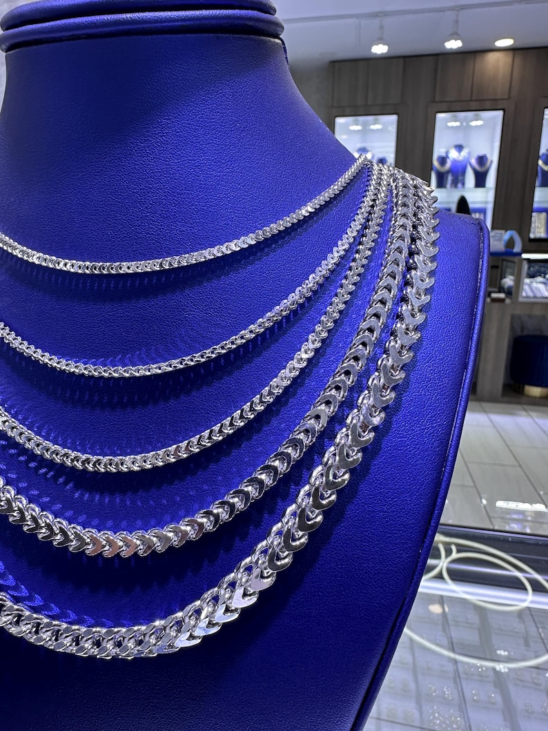 May include: A display of silver necklaces with various chain styles. The necklaces are arranged on a blue mannequin, showcasing different thicknesses and designs. The chains have a shiny, reflective surface.