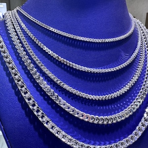 May include: A display of multiple silver chain necklaces with varying link designs. The necklaces are arranged on a blue velvet display, showcasing their different styles and sizes. The chains are reflective, catching the light and highlighting their intricate details.