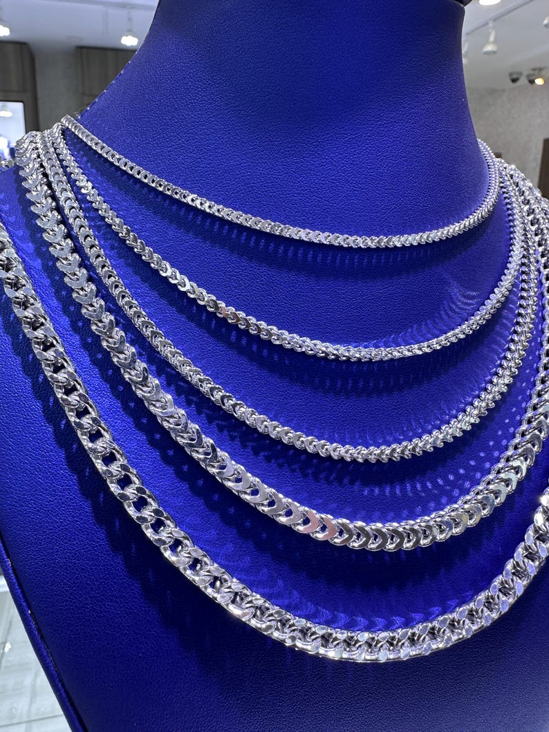 May include: A display of multiple silver chain necklaces with varying link designs. The necklaces are arranged on a blue velvet display, showcasing their different styles and sizes. The chains are reflective, catching the light and highlighting their intricate details.