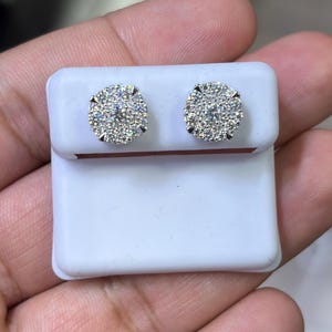 May include: Two round, cluster-style stud earrings with a sparkling, diamond-like appearance. The earrings are set on a white display card, showcasing their design. The studs are small and likely for pierced ears.