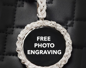 Puede incluir: Colgante plateado con diseño circular, con centro negro con el texto "FREE PHOTO ENGRAVING". El colgante está adornado con numerosas piedras preciosas pequeñas y transparentes alrededor del borde y una fianza trenzada.
