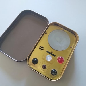 Könnte beinhalten: Eine rechteckige Metallbox im Vintage-Stil in Goldfarbe mit offenem Deckel. Die Box hat ein kreisförmiges Glaselement, verschiedene farbige Edelsteine und kleine Zifferblätter. Der Deckel ist dunkelbraun.