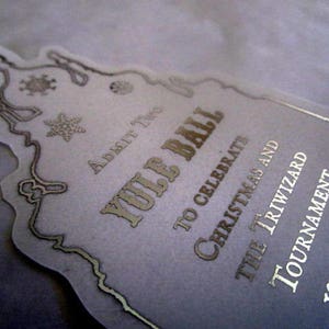 May include: A white invitation with silver lettering for a Yule Ball. The invitation features a stylized illustration of a castle with a snowflake and the text "Yule Ball", "To Celebrate Christmas and The Triwizard Tournament", and "19:00 Hours".
