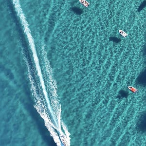 May include: Aerial view of a turquoise ocean with several boats. A speedboat leaves a white wake as it moves across the water. Other small boats are scattered throughout the scene, creating a sense of activity and leisure.