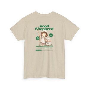 Könnte beinhalten: Beiges T-Shirt mit einem grünen Aufdruck eines Hirten und eines Schafs, mit den Worten "Good Shepherd". Das T-Shirt enthält auch den Text "JESUS SHEPHERD SAVIOR KING" und "JESUS SAID". Religiöses Thema.