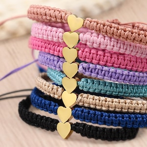 May include: A stack of colorful macrame bracelets in shades of pink, purple, blue, and tan. Each bracelet features a gold heart charm. The bracelets are handmade and have adjustable cords.