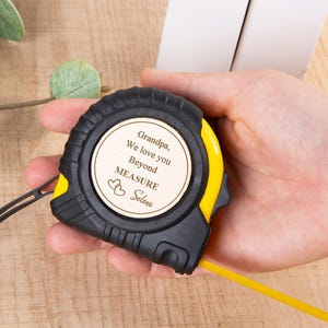 Puede incluir: Cinta métrica negra y amarilla con una inserción de madera personalizada. La inserción dice "Grandpa, We love you Beyond MEASURE Selena" con dos símbolos de corazón. La cinta métrica se sostiene en una mano.